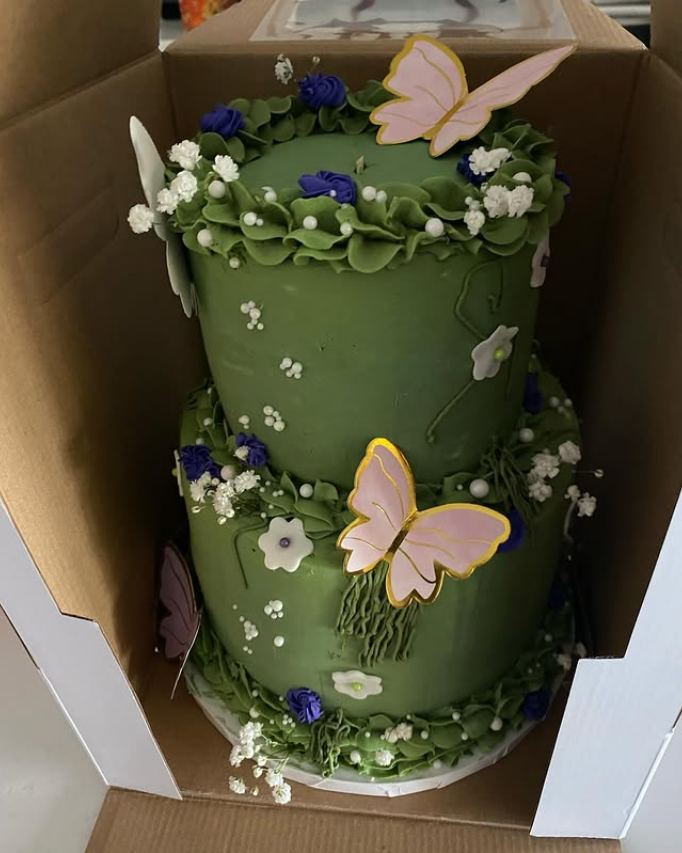 Two-tier green cake with purple and white buttercream flowers, baby’s breath, and pink and gold butterflies, in a cake box.